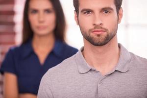 Young and successful. Confident young man looking at camera while woman standing behind him and keeping arms crossed photo