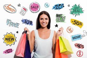 Customer choice. Cheerful young woman in dress carrying colorful shopping bags and looking at camera with colorful sketches upon her head photo