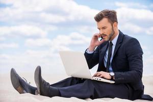 Staying in touch anytime and everywhere. Confident young man in formalwear working on laptop and talking on mobile phone while sitting on sand in desert photo