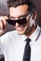 Feeling flirty. Handsome young man in shirt and tie adjusting his sunglasses and looking at camera with smile photo