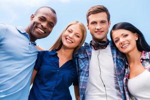 We are all good friends. Low angle view of four happy young people bonding and looking at camera with smile with blue sky in the background photo