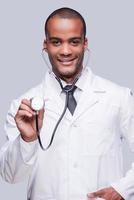 Examen médico. seguro médico africano estirando su estetoscopio y sonriendo mientras está de pie contra el fondo gris foto
