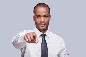 Te escojo a ti. un joven africano confiado con camisa y corbata apuntándote mientras te enfrentas a un fondo gris foto