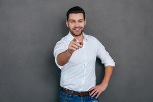 I choose you Handsome young man pointing you and smiling while standing against grey background photo