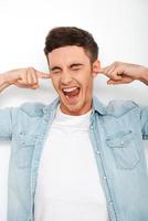 Stop it Frustrated young man holding fingers in ears and screaming while standing against white background photo