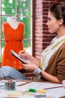 Young and full of creativity. Side view of beautiful young woman drawing fashion sketch and looking away while sitting at her working place with mannequin standing in the background photo