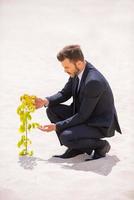 Growing up new business. Confident young businessman examining plant growing out of sand photo