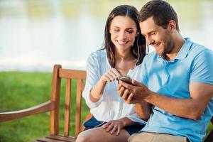 Enjoying favorite their music together. Beautiful young loving couple sitting on the bench together while woman pointing mobile phone and smiling photo