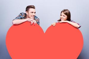 te amo hermosa joven pareja amorosa sonriendo y apoyándose en una enorme tarjeta de San Valentín en forma de corazón y sonriendo mientras está de pie contra un fondo gris foto