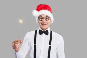 feliz año nuevo alegre joven nerd con corbata de moño y tirantes sosteniendo una bengala y sonriendo mientras se enfrenta a un fondo gris foto