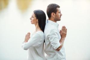 encontrar la armonía dentro de sí mismos. vista lateral de una hermosa pareja joven vestida de blanco meditando juntos al aire libre mientras están de pie espalda con espalda foto