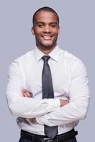 Young and successful. Confident young African man in shirt and tie keeping arms crossed and smiling while standing against grey background photo