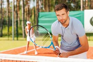 Concentrated on game. Handsome young man holding tennis racket and looking away while standing on tennis court and with woman in the background photo