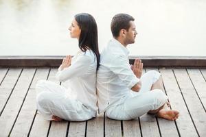 In peace with universe. Side view of beautiful young couple in white clothing meditating outdoors together while sitting back to back in lotus position