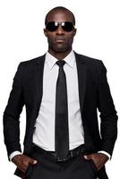 Cool and confident. Serious young African man in formalwear and sunglasses holding hands in pockets and looking at camera while standing isolated on white background photo