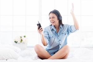 It is my favorite song Happy young woman in headphones listening to MP3 player and gesturing while sitting in bed photo