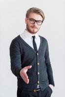 Welcome on board Handsome young man in glasses stretching out hand for shaking while standing against grey background photo