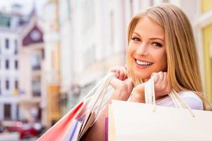 feliz chica adicta a las compras. hermosa joven mujer alegre sosteniendo bolsas de compras y mirando a la cámara mientras está de pie al aire libre foto