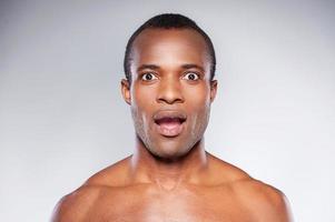 qué retrato sorpresa de un hombre africano sorprendido y sin camisa mirando a la cámara y manteniendo la boca abierta mientras está aislado en un fondo gris foto