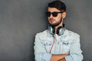 Confident and stylish. Young man in headphones keeping arms crossed and looking away while standing against grey background photo