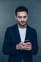 Important massage. Confident young man holding mobile phone and typingwhile standing against grey background photo