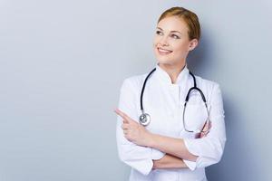 Look over there Confident female doctor in white uniform looking away and pointing while standing against grey background photo