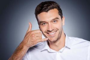 joven y exitoso. joven feliz con camisa blanca haciendo gestos con el teléfono móvil cerca de la cara y sonriendo mientras se enfrenta a un fondo gris foto