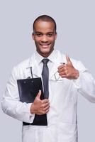 Good news. Cheerful African doctor showing his thumb up and smiling while standing against grey background photo