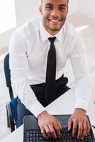 Businessman at work. Top view of confident young African man in shirt and working on laptop and smiling while sitting at his working place photo