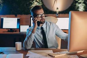 Confident young man working on computer and talking on mobile phone while staying late in the office photo
