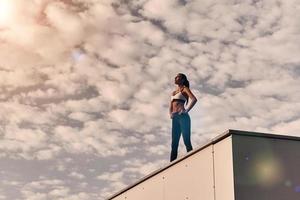 su cuerpo se ganó un descanso. mujer joven moderna en ropa deportiva relajándose mientras está de pie en el techo al aire libre foto