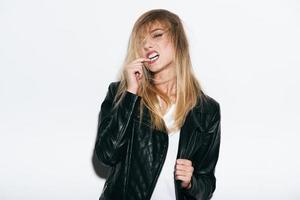 Cool and free. Funky young woman in leather coat holding finger in mouth while standing against white background photo