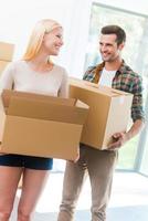 Happy to start a new life together. Happy young couple holding a cardboard box and looking at each other while other carton boxes laying on background photo