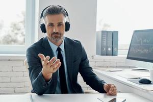 hombre de negocios confiado en auriculares gesticulando mientras está sentado en su lugar de trabajo foto