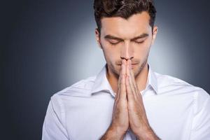 Man praying. Concentrated young man in white shirt holding hands clasped near face while standing against grey background photo