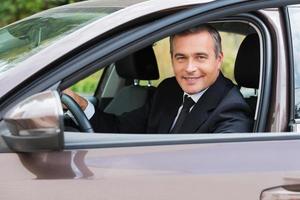 este coche es simplemente un hombre maduro, alegre y perfecto con ropa formal mirando el coche y sonriendo mientras está sentado en el coche foto