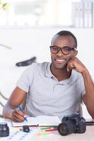 I love my job Cheerful young African man in casual wear writing something in note pad and looking at camera while sitting at his working place photo