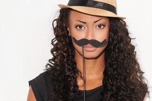 What do you think about my mustache Portrait of playful young African woman in funky hat holding fake mustache on her face and looking at camera while standing against white background photo