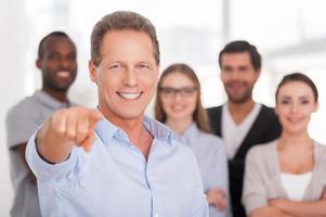 I choose you Confident mature man pointing you and smiling while group of people in casual wear standing on background photo