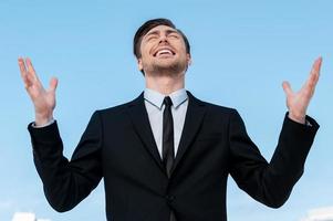 Thanks to God Handsome young man in suit rising hands up and smiling while standing against blue sky photo