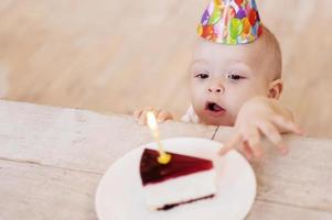 My first birthday cake Top view of cute little baby in party hat stretching hand to the plate with cake and keeping mouth open photo