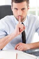 Keep silence Confident young man in formalwear holding finger on mouth and looking at camera while sitting at his working place photo