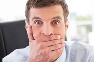 No way Shocked young man in formalwear covering mouth with hand and looking at camera while sitting at his working place photo