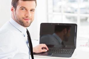 How may I help you Handsome young man in formalwear working on laptop and looking over shoulder while sitting at his working place photo