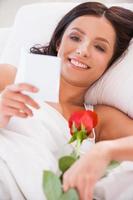 Message from boyfriend. Close up image  of beautiful young woman lying in bed with red rose and reading letter photo