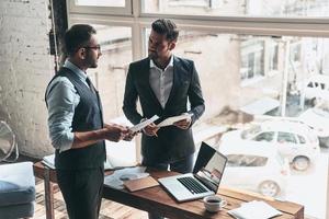 Going over details. Top view of two young modern men in formalwear working together while standing indoors photo