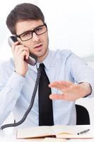 no ahora un joven enojado con camisa y corbata hablando por teléfono móvil y gesticulando mientras está sentado en su lugar de trabajo foto