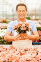 This flower is yours Handsome mature man in apron stretching out a potted plant and smiling while standing in a greenhouse photo