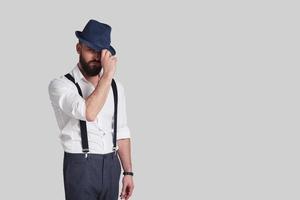 Looks like gangster. Handsome young man in suspenders adjusting his hat and looking at camera while standing against grey background