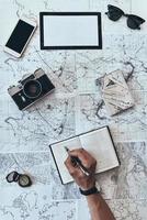 Planning travel. Close up top view of man writing something down in diary with sunglasses, photo camera, compass, passport and smart phone lying on map around